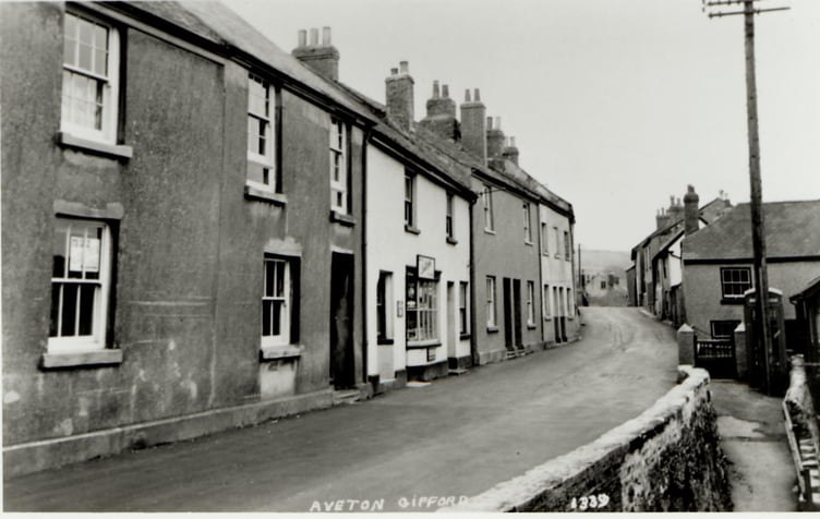 Telephone box on right hand side of Fore Street, Aveton Gifford. Provision store on left. Brick wall in front of telephone box. Around 1910.