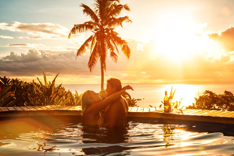 Photo of a young couple enjoying a romantic sunset in Fiji as they cool off in the pool with glasses of wine; kissing.